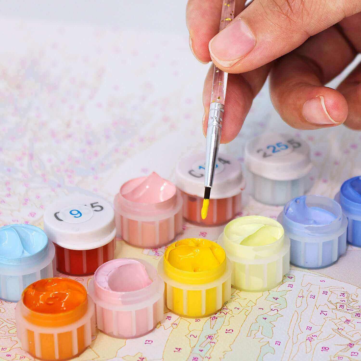 Hand holding a brush over vibrant paint pots for a paint by numbers kit, ready to create art.